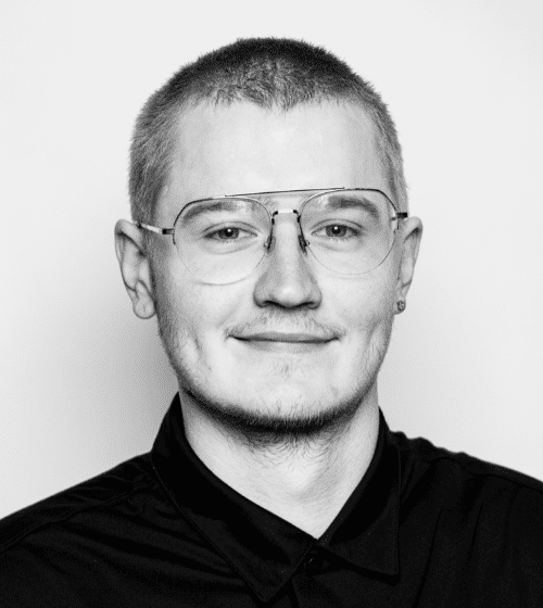 Black and white portrait of a person with short hair, glasses, a faint mustache, and an earring, wearing a dark shirt and smiling slightly.