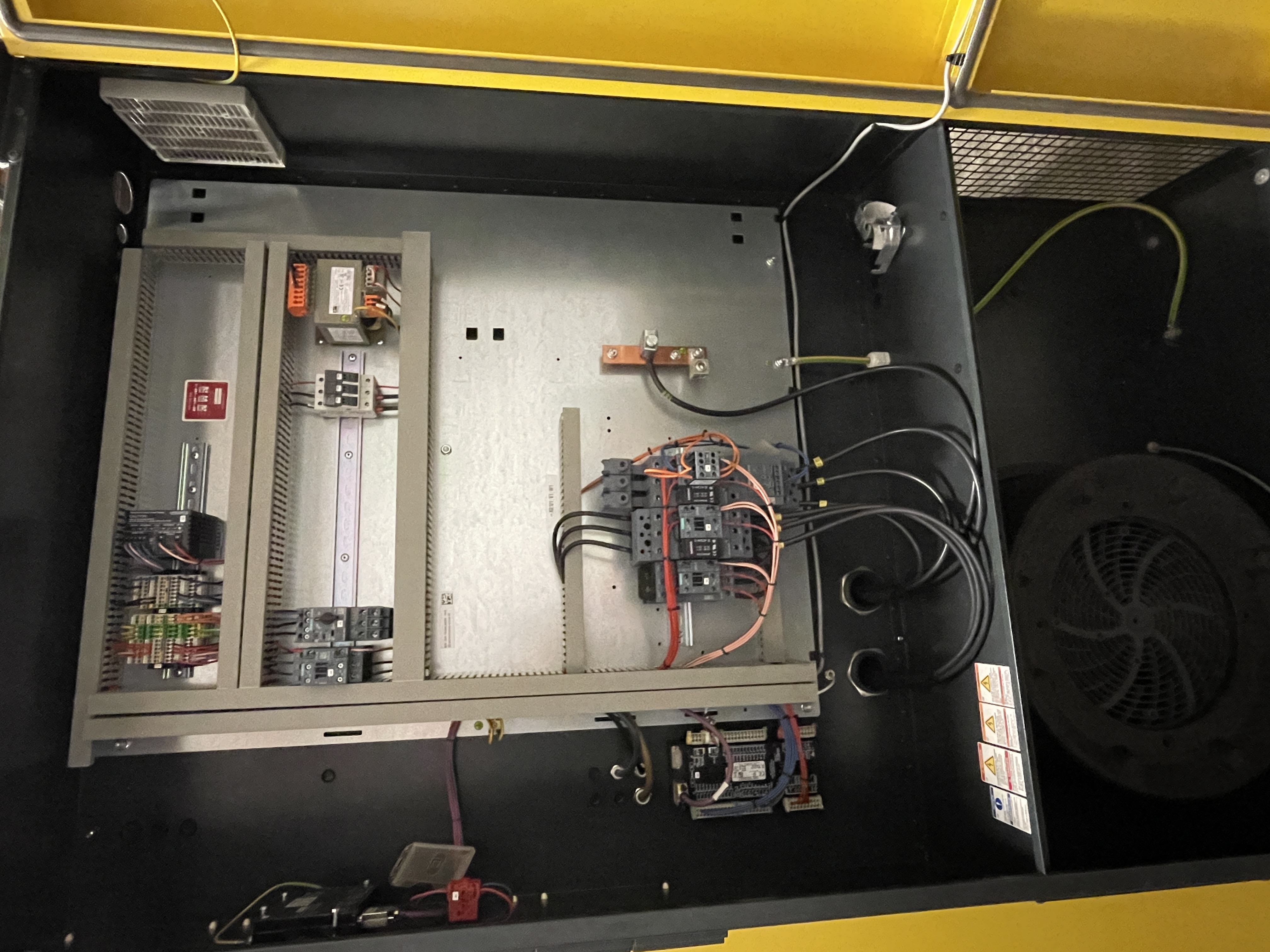 An open electrical panel with various wires, circuit breakers, and components mounted inside, enclosed by a yellow frame.