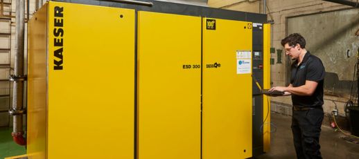 A person stands next to a large yellow industrial machine labeled "KAESER ESD 300," using a laptop.