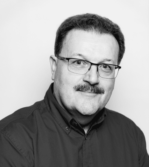 A man with short dark hair, mustache, and glasses, wearing a dark button-up shirt, smiles at the camera. Black and white image.