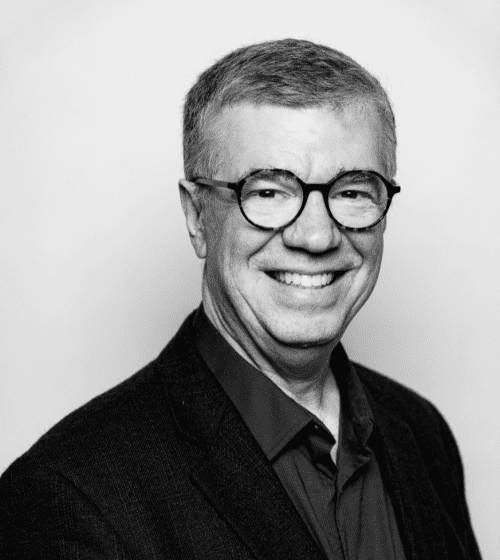 Black and white photo of a smiling man with short hair, wearing round glasses, a dark blazer, and a button-up shirt, standing against a plain background.