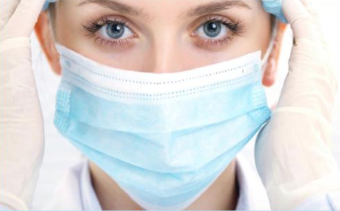 A person with blue eyes wears a blue surgical mask, cap, and gloves, adjusting their mask with both hands.