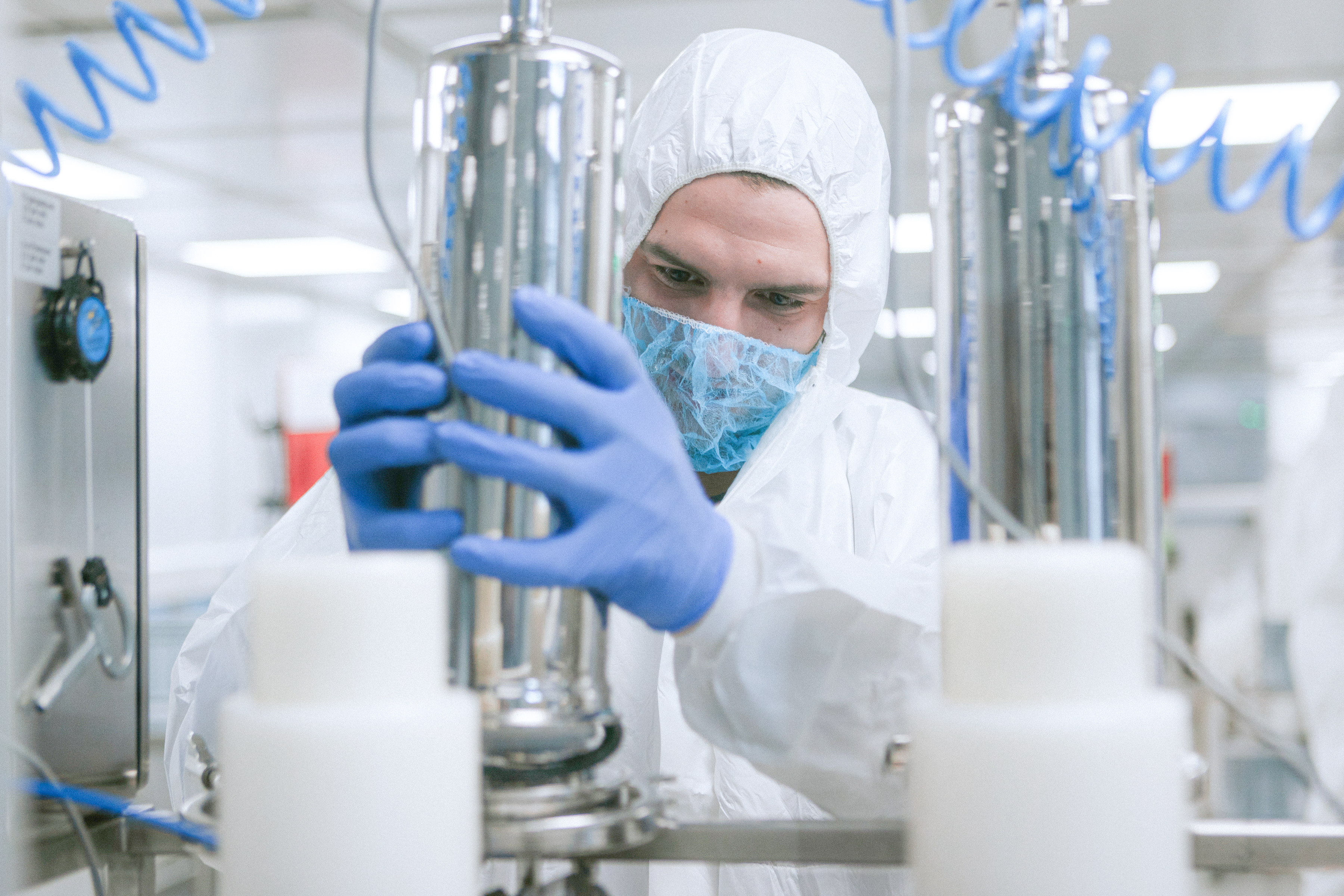 A person wearing protective gear, including gloves, a hairnet, a face mask, and a white suit, is handling silver cylindrical equipment in a clean laboratory or industrial environment.