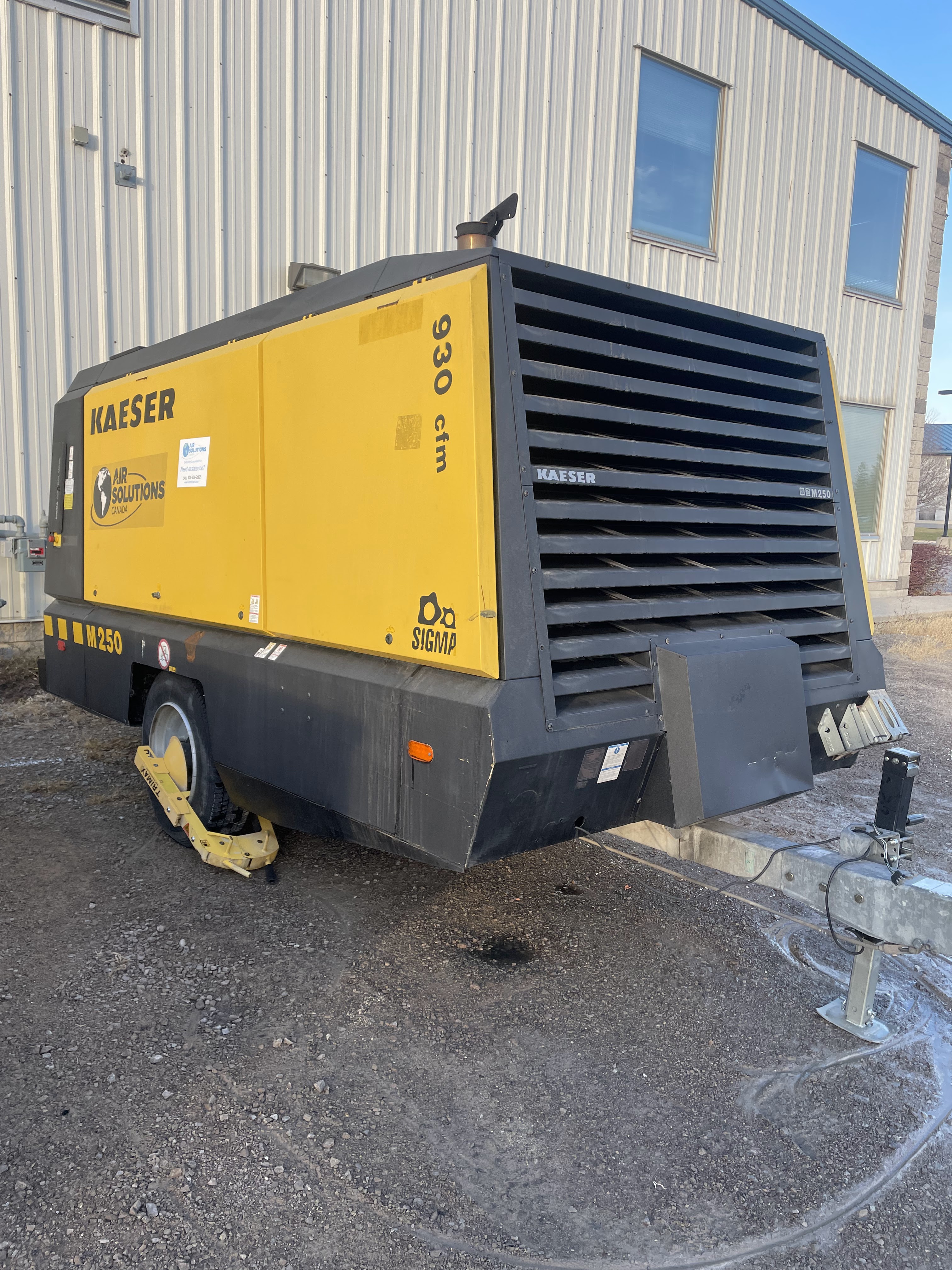 A yellow and black Kaeser M250 industrial air compressor is parked on gravel beside a metal building, with a wheel clamp attached to one tire.