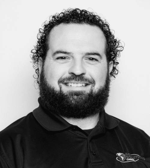 A man with curly hair and a beard, smiling, wearing a collared shirt with a logo on the right side. Black and white photo.