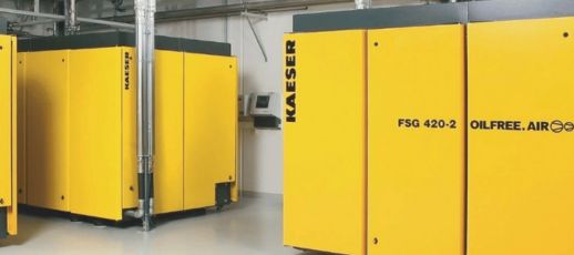 Yellow industrial air compressors labeled "KAESER" and "OILFREE.AIR" are positioned in a clean, modern facility with grey flooring and silver piping in the background.