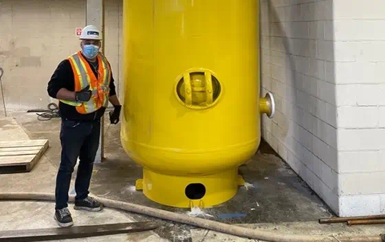 A person wearing a safety vest, mask, and gloves stands next to a large yellow industrial tank inside a facility.
