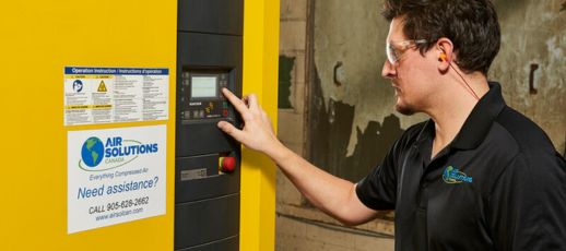 A man in safety glasses operates a machine with a touch screen. He's wearing a black shirt with a company logo. A yellow panel has instruction labels and a contact sign for assistance.