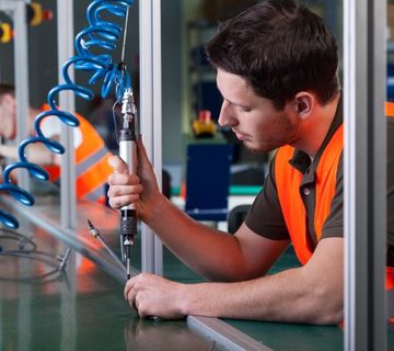 A worker in an orange safety vest uses a power tool on an assembly line.