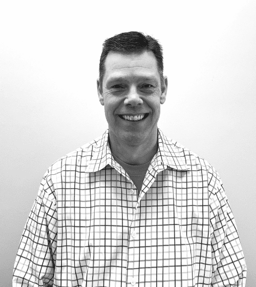 Black and white photo of a man wearing a checkered shirt, smiling, and standing in front of a plain, light-colored background.